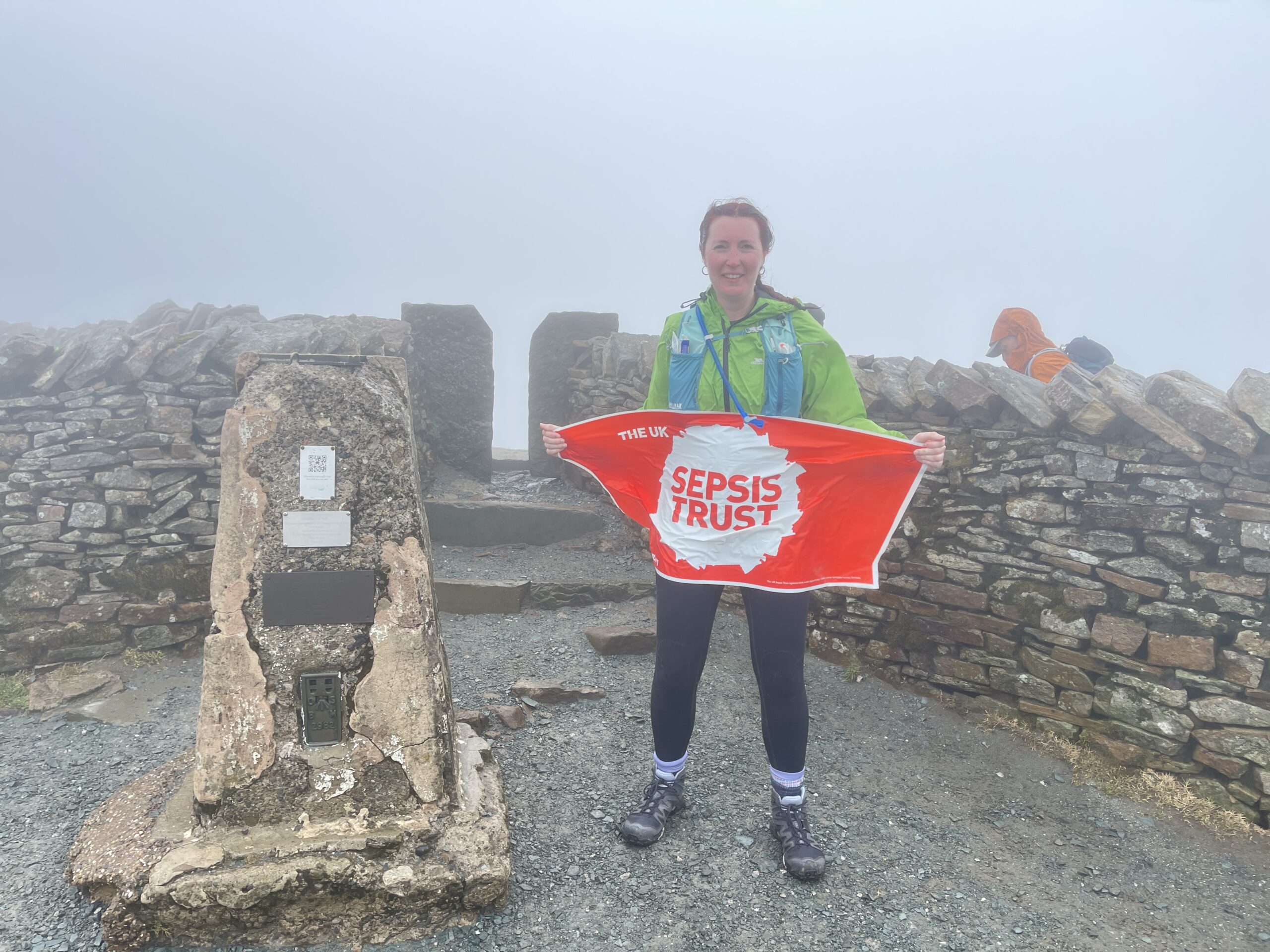 A woman stands on the top of a hill, in the fog, holding a banner that has the UKST logo on.