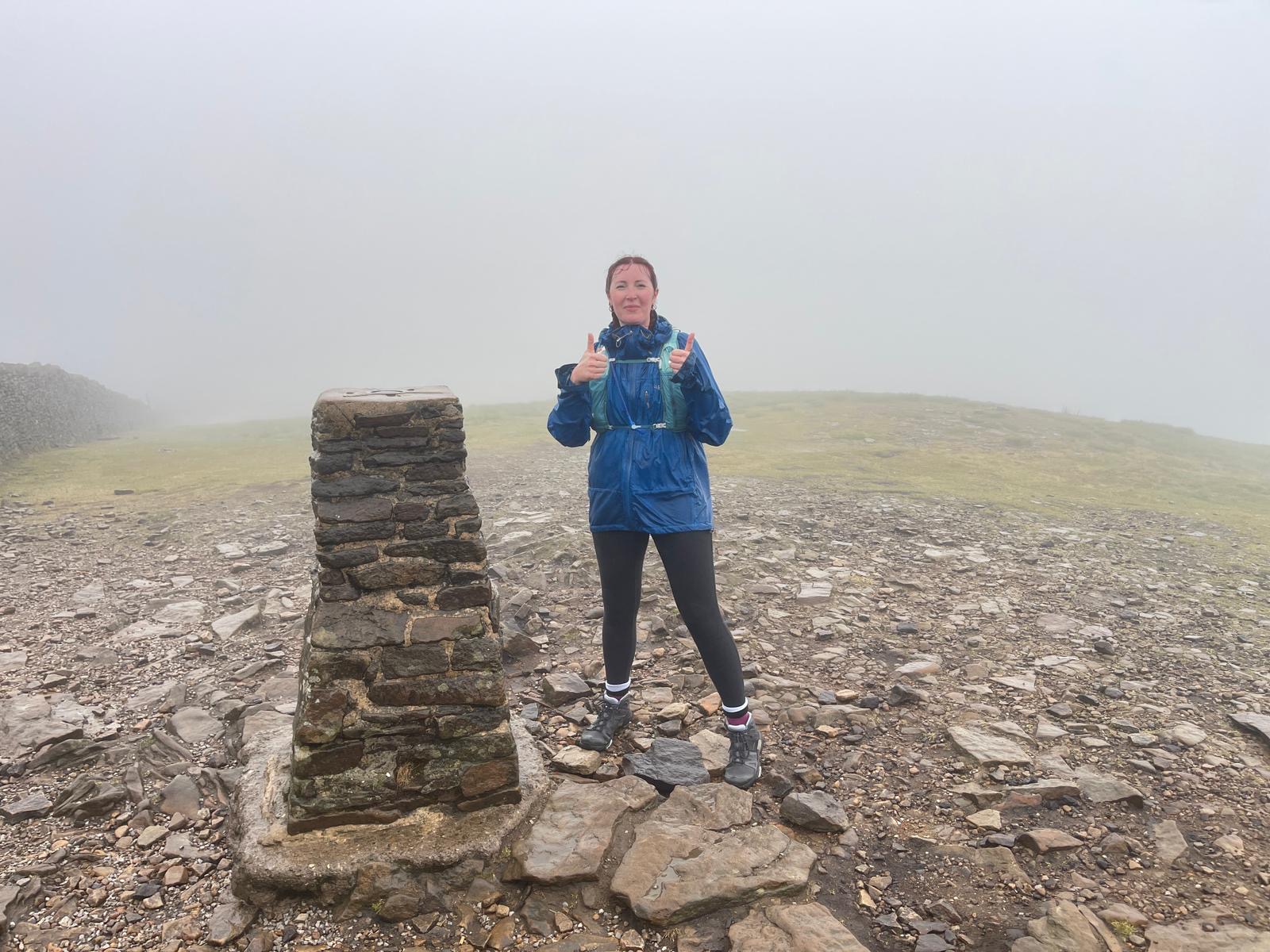 A woman stands on top of a hill in the Yorkshire Dales in the rain and wind, smiling.