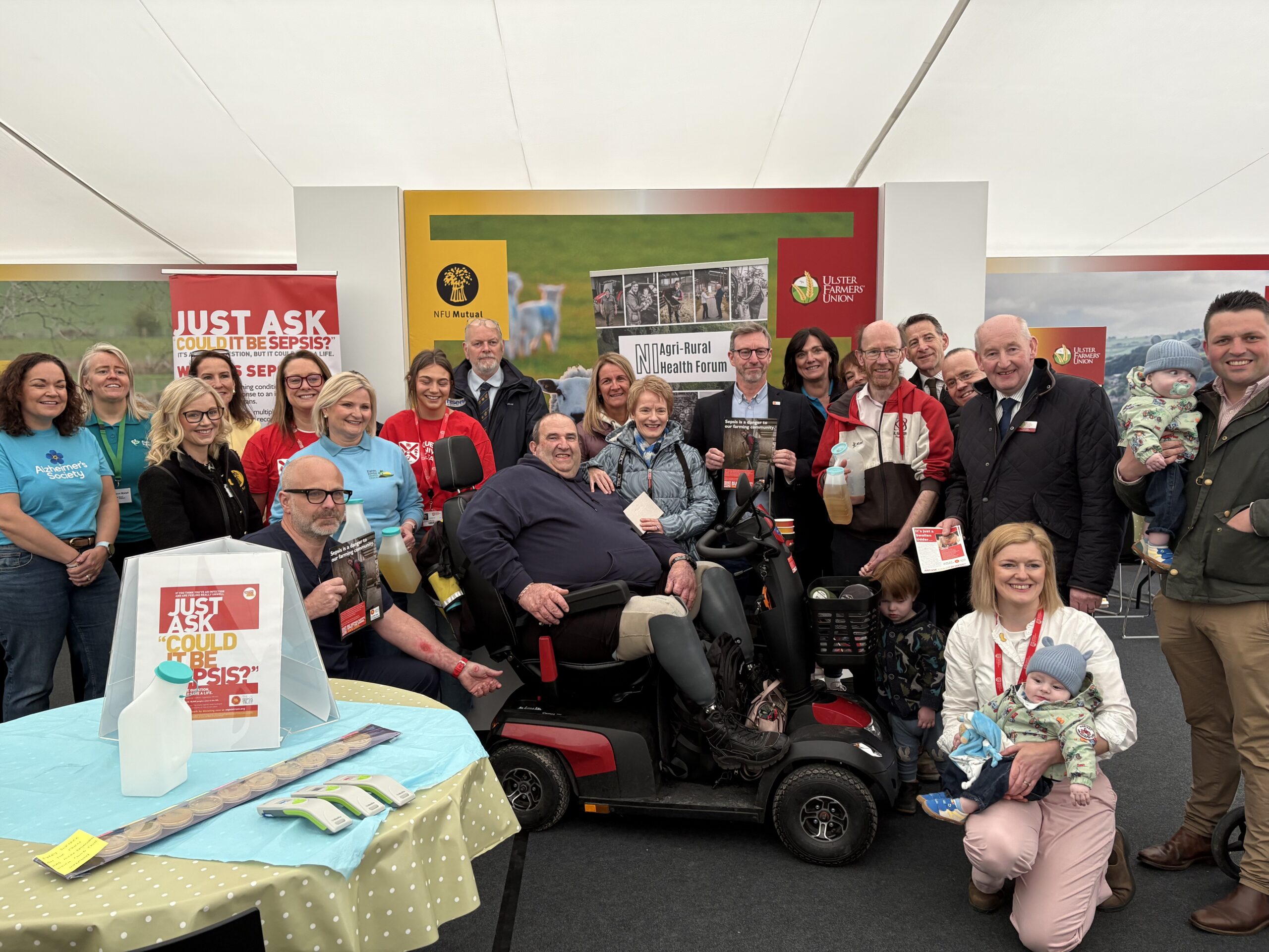 IMG_2306 Large group gathered at a Balmoral Show exhibition stand for a sepsis awareness campaign, with signage reading “Just ask: could it be sepsis?” and participants holding information materials focused on farming communities.
