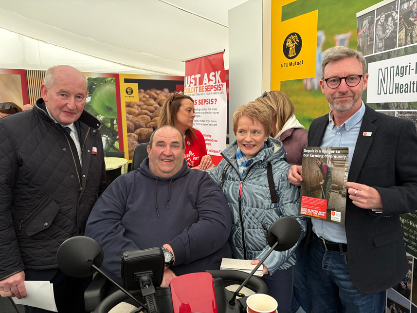 Group gathered at a Balmoral Show stand launching a sepsis awareness campaign for farming communities, with campaign banners reading “Just ask: could it be sepsis?” and a leaflet highlighting the risks and warning signs of sepsis.