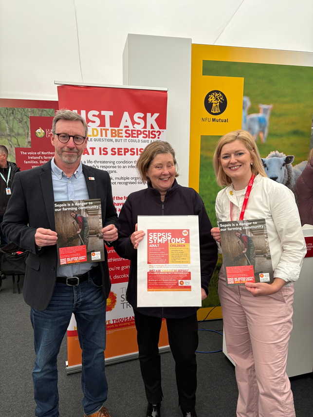 Three people at the Balmoral Show display sepsis awareness materials, including a symptoms poster and leaflets stating “Sepsis is a danger to our farming community,” promoting early recognition and urgent action.