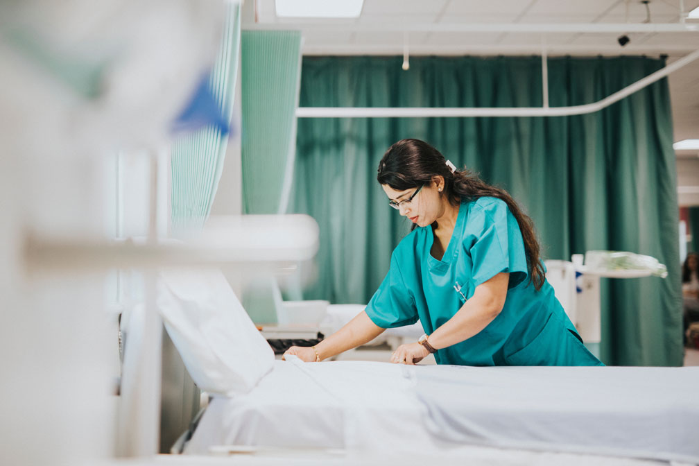 Hospital corridor with a folded patient trolley in the foreground and several healthcare staff in scrubs working in the distance. Bright overhead lighting reflects off the polished floor, and stainless steel doors line both sides of the corridor.