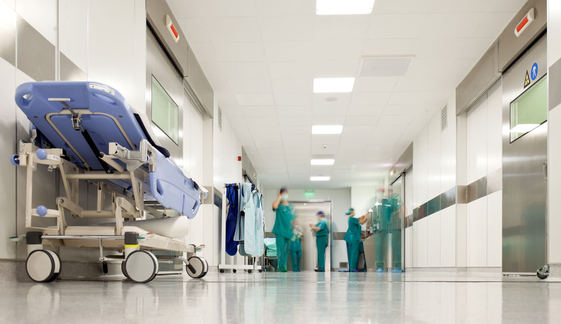 Hospital corridor with a folded patient trolley in the foreground and several healthcare staff in scrubs working in the distance. Bright overhead lighting reflects off the polished floor, and stainless steel doors line both sides of the corridor.