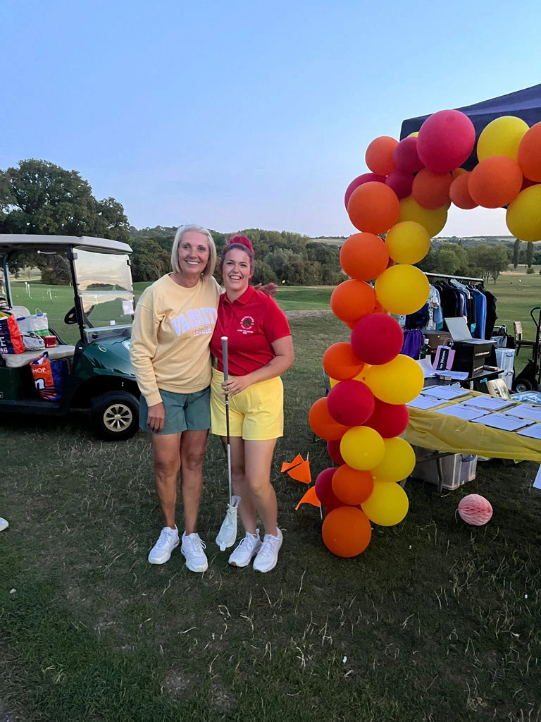 Two people stand together on the golf course beside a red, orange and yellow balloon arch at a charity golf day. A fundraising table, golf buggy and the course landscape can be seen in the background.
