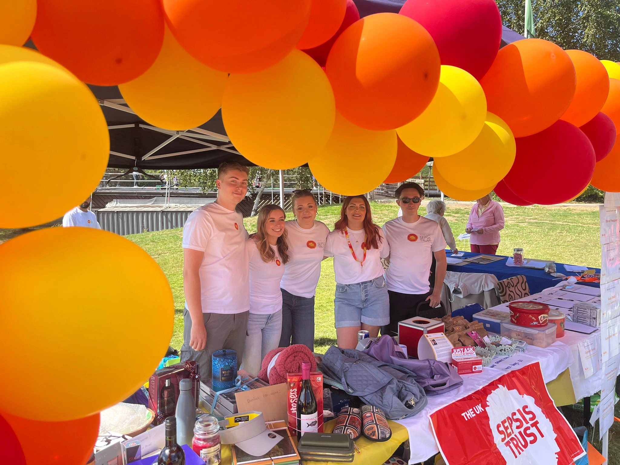 Five volunteers pose behind a fundraising and information stall decorated with large red, orange and yellow balloons during a charity golf day. The table is covered with raffle prizes, leaflets and charity materials.