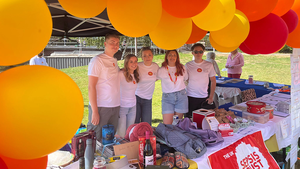 Golf events page Five volunteers pose behind a fundraising and information stall decorated with large red, orange and yellow balloons during a charity golf day. The table is covered with raffle prizes, leaflets and charity materials.