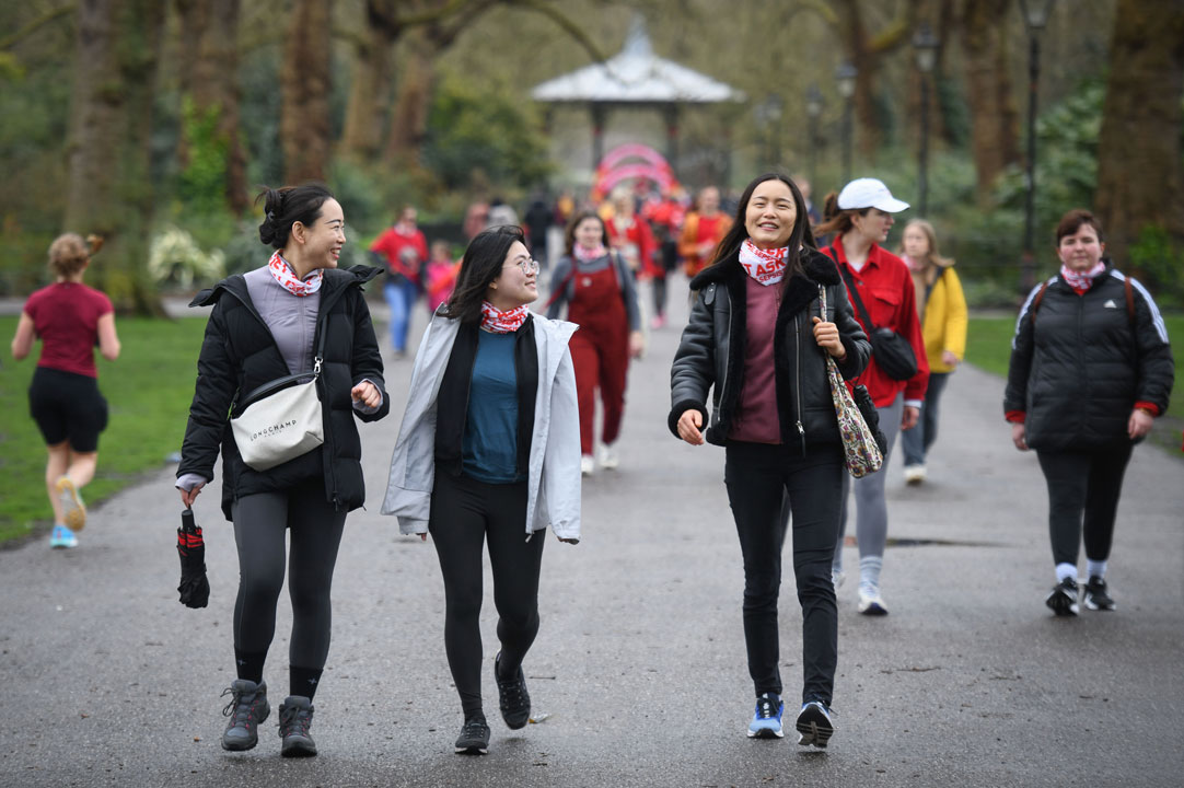 A group of people walk along a wide path in a park lined with tall trees. Three people in the foreground wear coats, leggings, and patterned neck scarves, while other participants walk and jog behind them. A gazebo is visible in the blurred background, and the overcast weather gives the scene a cool, early-spring atmosphere.