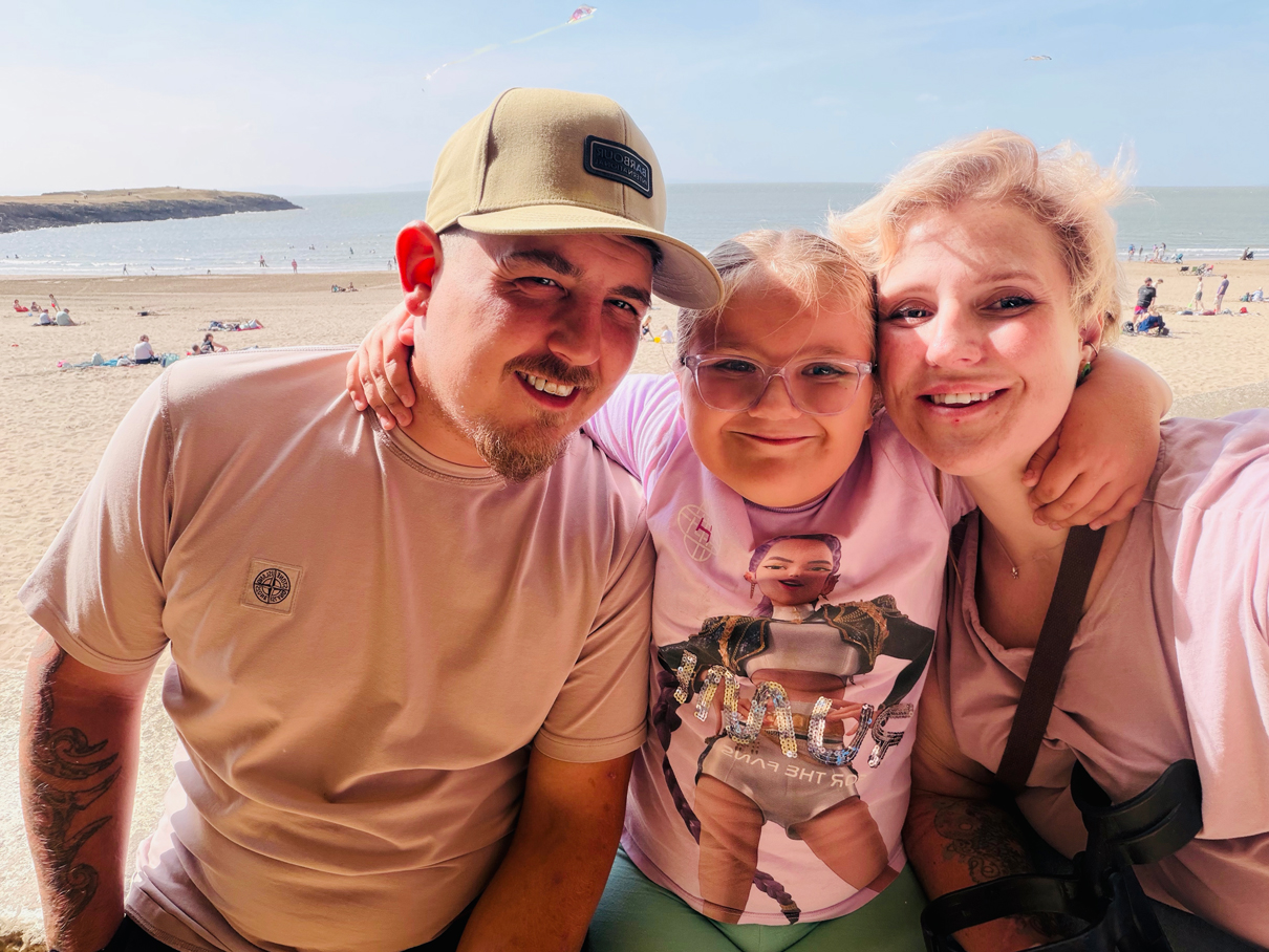 Three people pose together for a selfie at a sunny beach, with the sea, sandy shoreline, and distant headland visible in the background.