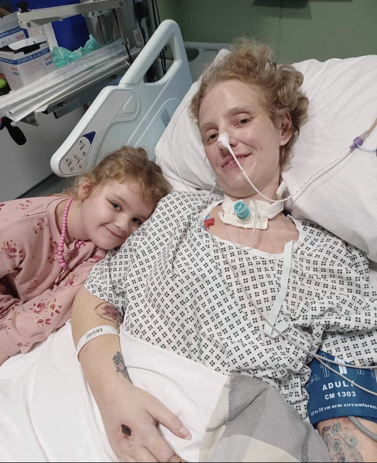 A hospital patient resting in bed with medical tubes and monitoring equipment is joined by a young child leaning against their shoulder in a hospital room.