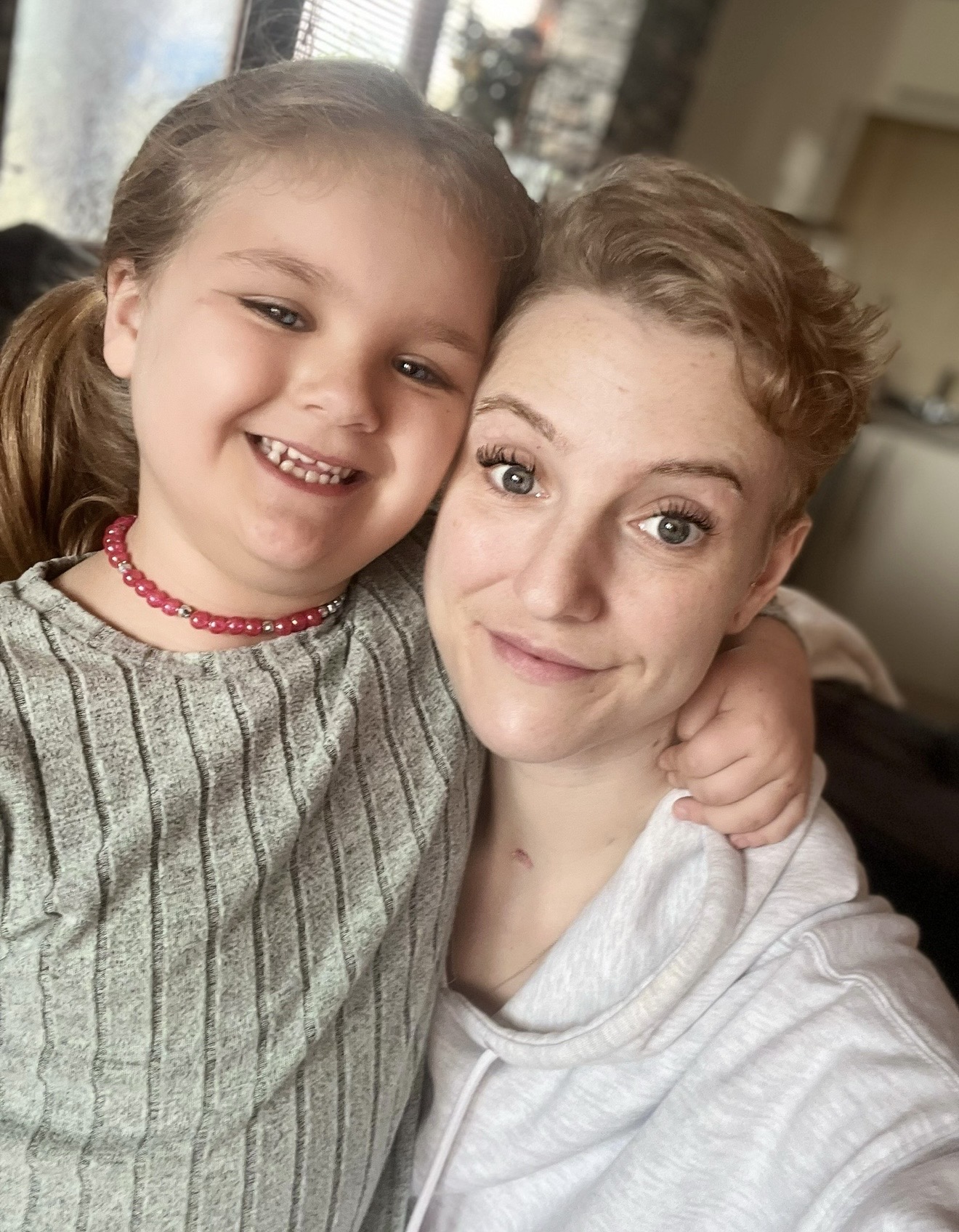 A child and an adult sit close together indoors for a portrait photo, with the child’s arm around the adult’s shoulders and a softly blurred home setting in the background.